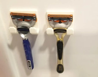 May include: Two razor holders with razors attached. The holders are white plastic and are mounted on a white wall. The razors are both Gillette brand, one with a blue handle and one with a gold handle.