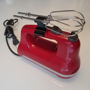 Puede incluir: Una batidora de mano roja con un accesorio de batidor plateado. La batidora tiene un cable negro y un dial de control de velocidad plateado.