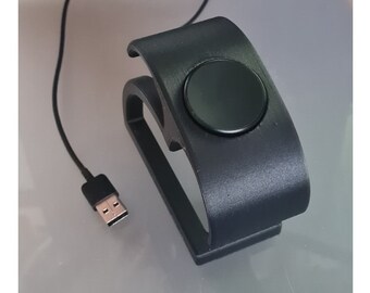 Puede incluir: Un soporte de carga negro con una plataforma de carga circular para un reloj inteligente. El soporte tiene un cable USB.