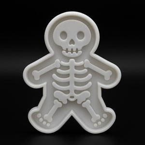 Könnte beinhalten: Ein weißer Ausstecher in Form eines Skelett-Lebkuchenmanns. Der Ausstecher zeigt einen Totenkopf, einen Brustkorb und Knochen der Gliedmaßen. Der Hintergrund ist einfarbig schwarz.