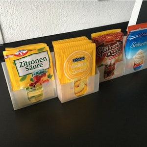 Puede incluir: Cuatro contenedores de plástico transparente con paquetes de mezcla de bebida en polvo de color amarillo, naranja y azul. Los paquetes tienen texto en alemán, incluyendo "Zitronen Säure", "Vanillin Zucker", "Glühwein Quick" y "Sahnesteif".
