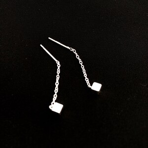 May include: A pair of silver threader earrings. Each earring features a delicate chain that ends with a small, square charm. The earrings are displayed against a black background.