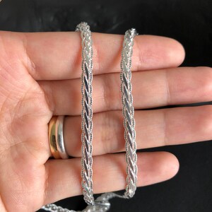 May include: A silver chain with a braided design. The chain is held in a hand, against a black background. The chain has a delicate, intricate pattern, and appears to be made of metal. The chain is a jewelry item.