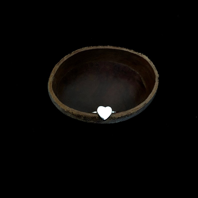 K&ouml;nnte beinhalten: Ein silberner Ring mit herzf&ouml;rmigem Design. Der Ring ist vor einem dunklen Hintergrund platziert, mit einem braunen, schalenf&ouml;rmigen Objekt im Hintergrund. Das Herz ist glatt und poliert.
