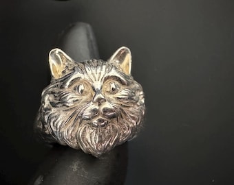 Könnte beinhalten: Ein silberner Ring mit einem detaillierten Katzenkopf-Design. Der Ring zeigt die Merkmale der Katze, einschließlich Ohren, Augen, Nase und Schnurrhaare. Der Ring ist vor einem dunklen Hintergrund platziert.