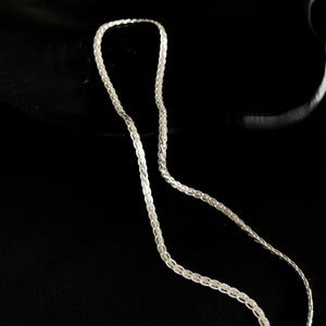 May include: A silver chain necklace with a detailed, woven pattern. The necklace is shown against a dark backdrop, emphasising its intricate design and metallic shine. The chain is composed of small, interconnected links.