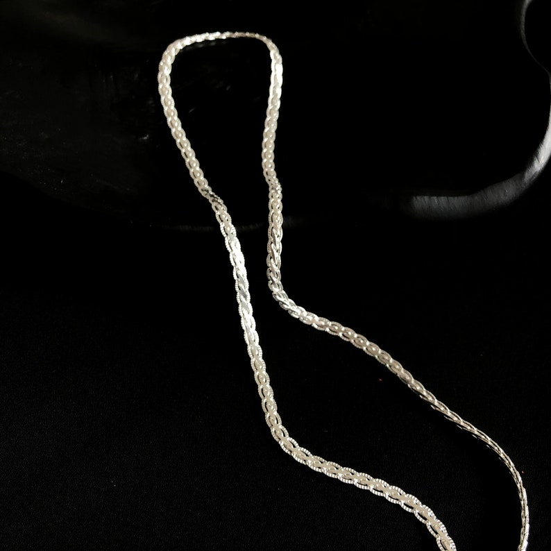 May include: A silver chain necklace with a detailed, woven pattern. The necklace is shown against a dark backdrop, emphasising its intricate design and metallic shine. The chain is composed of small, interconnected links.