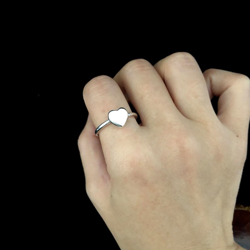 K&ouml;nnte beinhalten: Ein silberner Ring mit herzf&ouml;rmigem Design. Der Ring wird an einem Finger getragen, wobei das herzf&ouml;rmige Element deutlich sichtbar ist. Der Ringreif ist glatt und poliert und reflektiert das Licht. Der Hintergrund ist schwarz.