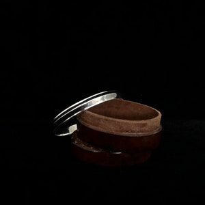 May include: A silver bangle bracelet with a black stripe rests on a brown leather container. The bracelet is highly reflective, and the container has a textured surface. The background is solid black.