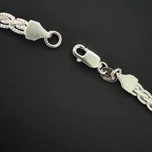 May include: Close-up of a silver chain necklace with a textured, woven design. The necklace is unclasped, showing the clasp and ring. The background is a solid black.