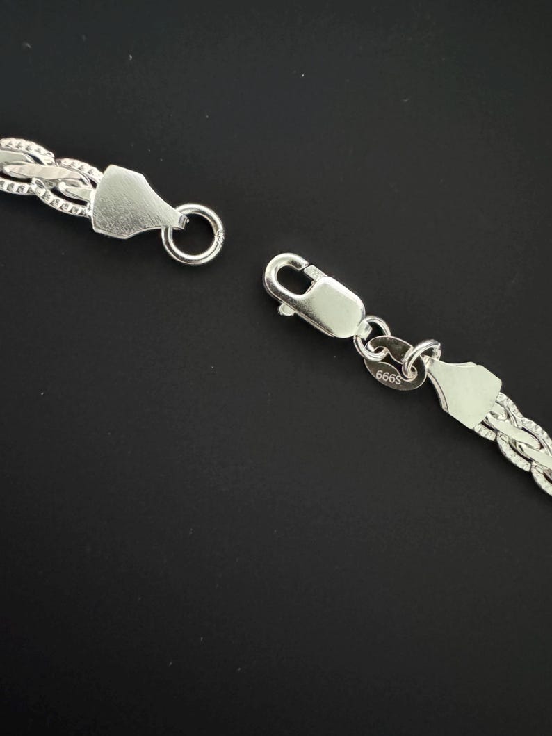 May include: Close-up of a silver chain necklace with a textured, woven design. The necklace is unclasped, showing the clasp and ring. The background is a solid black.