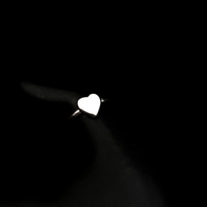 K&ouml;nnte beinhalten: Ein silberner Ring mit herzf&ouml;rmigem Design. Das Herz ist glatt und poliert und reflektiert das Licht vor einem schwarzen Hintergrund. Der Ring ist schmal und schlicht und erg&auml;nzt die Herzform.