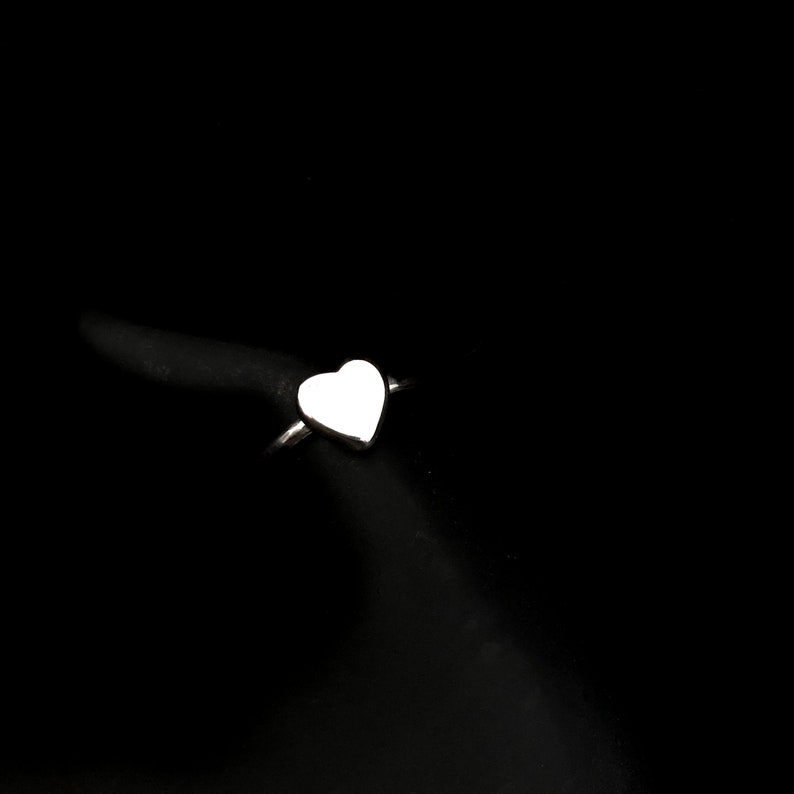 K&ouml;nnte beinhalten: Ein silberner Ring mit herzf&ouml;rmigem Design. Das Herz ist glatt und poliert und reflektiert das Licht vor einem schwarzen Hintergrund. Der Ring ist schmal und schlicht und erg&auml;nzt die Herzform.