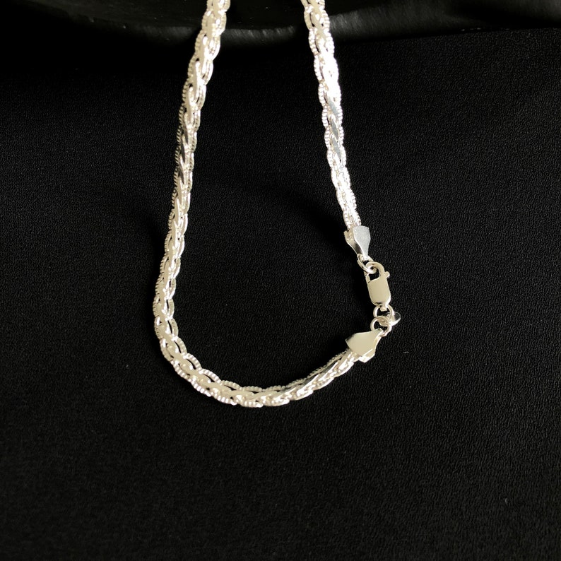 May include: A silver chain necklace with a woven pattern. The necklace features a lobster clasp fastening. The jewellery is shown against a black backdrop.