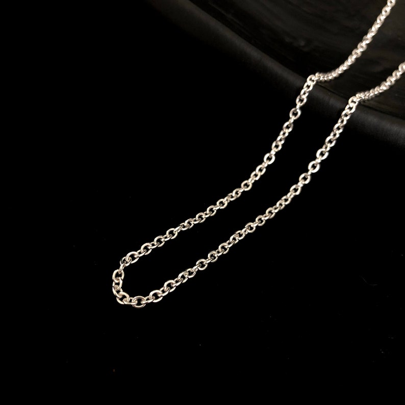 May include: A silver-colored chain necklace with small, round links. The chain is displayed against a black background, highlighting its reflective surface and delicate design. The necklace appears to be a simple, elegant piece of jewelry.