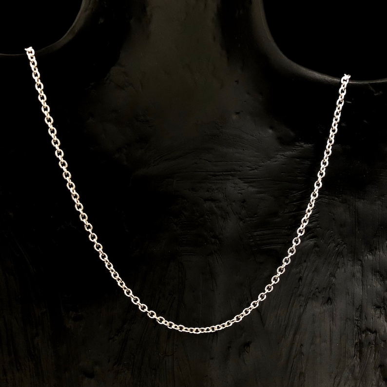 May include: A silver-colored chain necklace displayed on a black mannequin. The necklace is a delicate, simple design, with small, evenly spaced links. The chain is draped, showcasing its length and form. The background is a solid black.