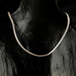 Puede incluir: Un collar de cadena de plata exhibido en un maniquí negro. La cadena tiene un diseño plano y estrechamente entrelazado, que refleja la luz. El collar es una joya, adecuada para diversas ocasiones.