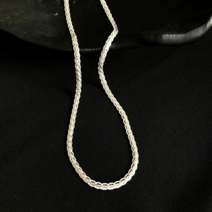 May include: A silver chain necklace with a delicate, woven design. The necklace is shown against a dark background, highlighting its intricate pattern and metallic sheen. The chain's links are closely connected, creating a smooth, continuous appearance.