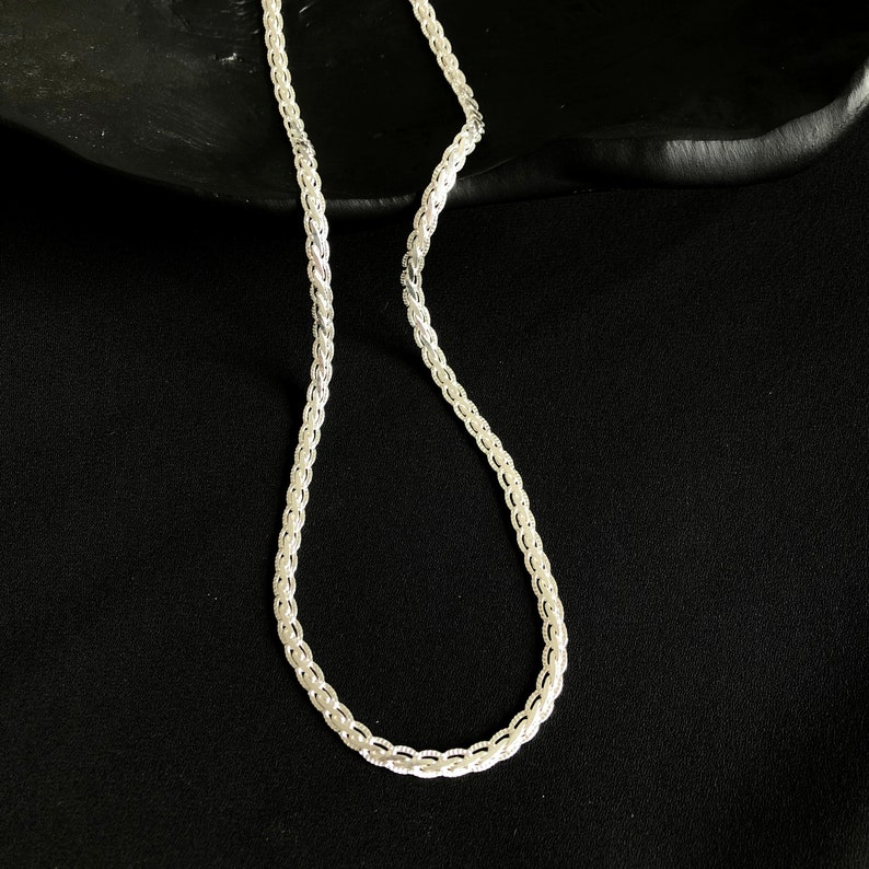 May include: A silver chain necklace with a delicate, woven design. The necklace is shown against a dark background, highlighting its intricate pattern and metallic sheen. The chain's links are closely connected, creating a smooth, continuous appearance.