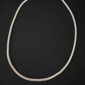 May include: A silver necklace featuring a woven chain design. The necklace is set against a black backdrop, accentuating its detailed pattern and metallic lustre. The chain is approximately 45 cm in length.