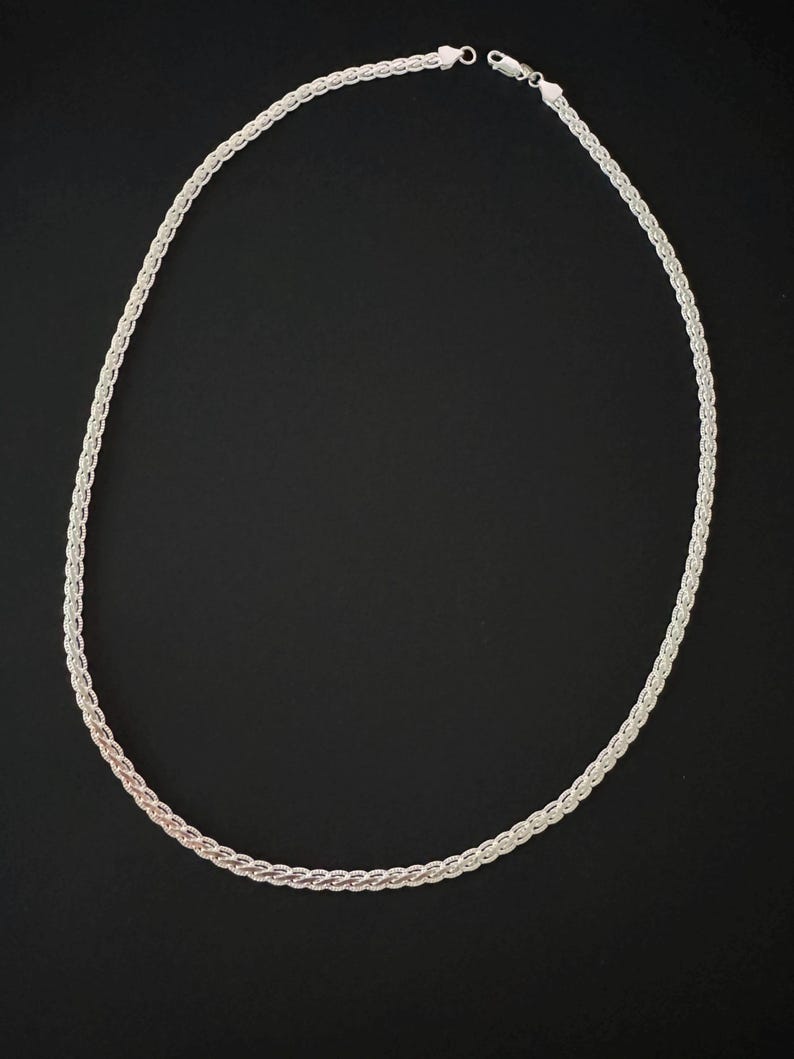 May include: A silver necklace featuring a woven chain design. The necklace is set against a black backdrop, accentuating its detailed pattern and metallic lustre. The chain is approximately 45 cm in length.