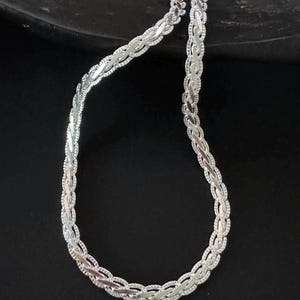 May include: A silver necklace featuring a braided chain design. The necklace is set against a dark backdrop, accentuating its detailed pattern and metallic lustre. The chain is composed of small, interconnected links.