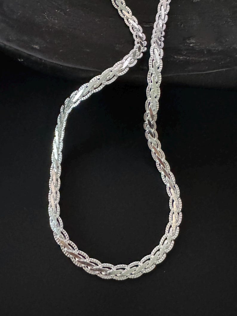May include: A silver necklace featuring a braided chain design. The necklace is set against a dark backdrop, accentuating its detailed pattern and metallic lustre. The chain is composed of small, interconnected links.