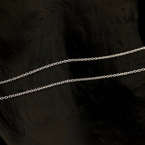 May include: A delicate silver chain necklace with a classic cable link design. The chain is displayed against a dark, textured background, highlighting its bright, reflective surface. This necklace is a simple yet elegant piece of jewelry.