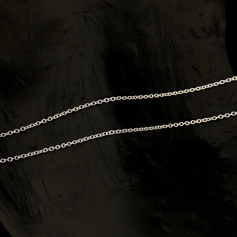 May include: A delicate silver chain necklace with a classic cable link design. The chain is displayed against a dark, textured background, highlighting its bright, reflective surface. This necklace is a simple yet elegant piece of jewelry.