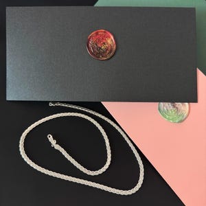 May include: A silver chain necklace with a textured design rests on a black surface. A black box with a red and gold wax seal is in the background. The background also includes pink and green surfaces.