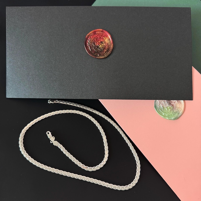 May include: A silver chain necklace with a textured design rests on a black surface. A black box with a red and gold wax seal is in the background. The background also includes pink and green surfaces.