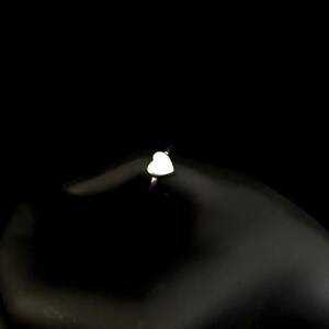 K&ouml;nnte beinhalten: Ein silberner Ring mit einem wei&szlig;en herzf&ouml;rmigen Design. Der Ring ist vor einem schwarzen Hintergrund platziert, der die zarten Details des Schmucks hervorhebt. Das Herz ist glatt und poliert und reflektiert das Licht.