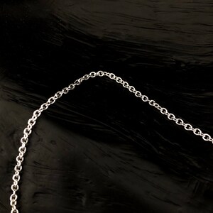 May include: A silver-colored chain necklace with small, round links. The chain is displayed against a dark, textured background, creating a contrast that highlights the necklace's design. The chain is curved, suggesting it is a necklace.