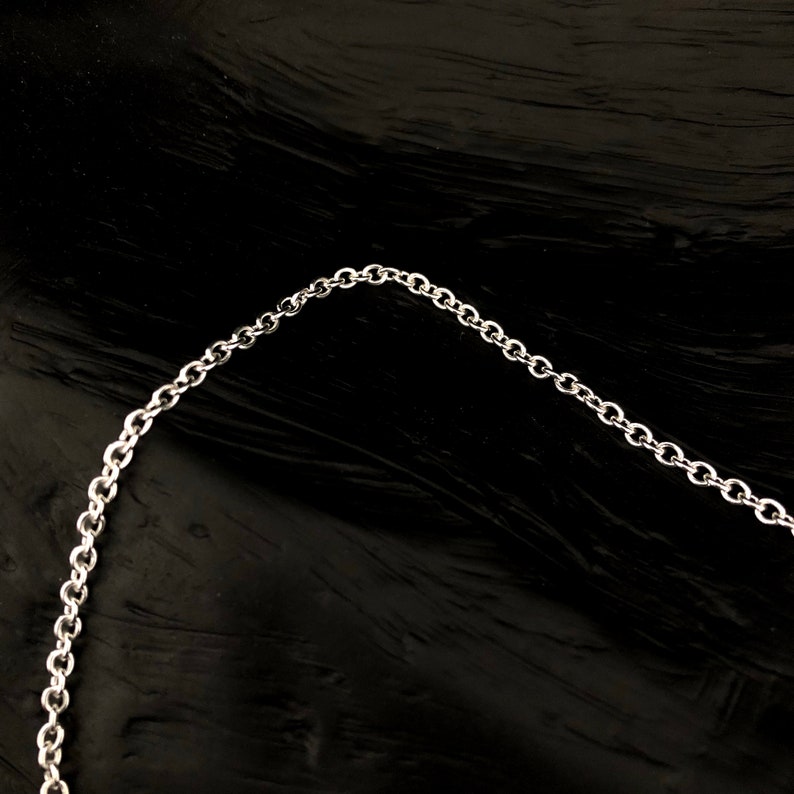 May include: A silver-colored chain necklace with small, round links. The chain is displayed against a dark, textured background, creating a contrast that highlights the necklace's design. The chain is curved, suggesting it is a necklace.