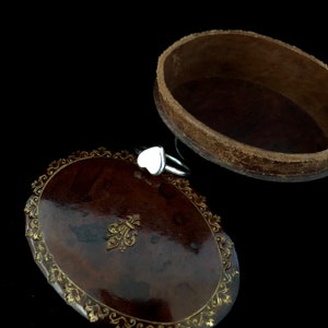 K&ouml;nnte beinhalten: Ein silberner Ring mit einem wei&szlig;en herzf&ouml;rmigen Stein liegt auf einer dunkelbraunen, ovalen Schmuckschatulle. Die Schatulle hat einen dekorativen goldfarbenen Rand und ein Fleur-de-Lis-Emblem. Die Schatulle ist ge&ouml;ffnet und gibt den Blick ins Innere frei.