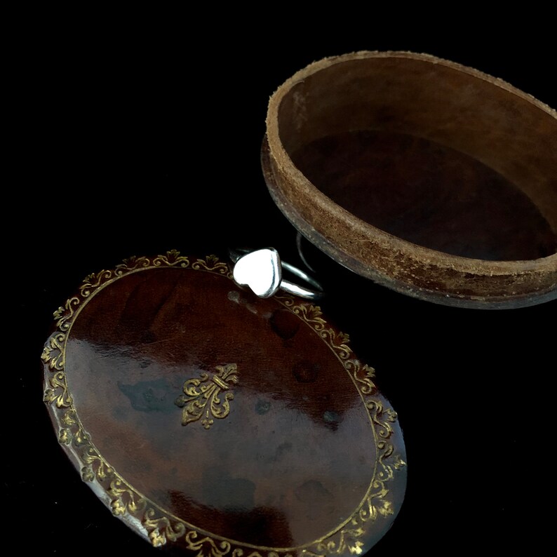 K&ouml;nnte beinhalten: Ein silberner Ring mit einem wei&szlig;en herzf&ouml;rmigen Stein liegt auf einer dunkelbraunen, ovalen Schmuckschatulle. Die Schatulle hat einen dekorativen goldfarbenen Rand und ein Fleur-de-Lis-Emblem. Die Schatulle ist ge&ouml;ffnet und gibt den Blick ins Innere frei.