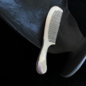 Könnte beinhalten: Ein silberfarbener Kamm mit gebogenem Griff und eng stehenden Zähnen. Der Kamm liegt auf einer dunklen, strukturierten Oberfläche, die mit seiner glänzenden Oberfläche kontrastiert. Der Griff hat ein glattes, abgerundetes Design.
