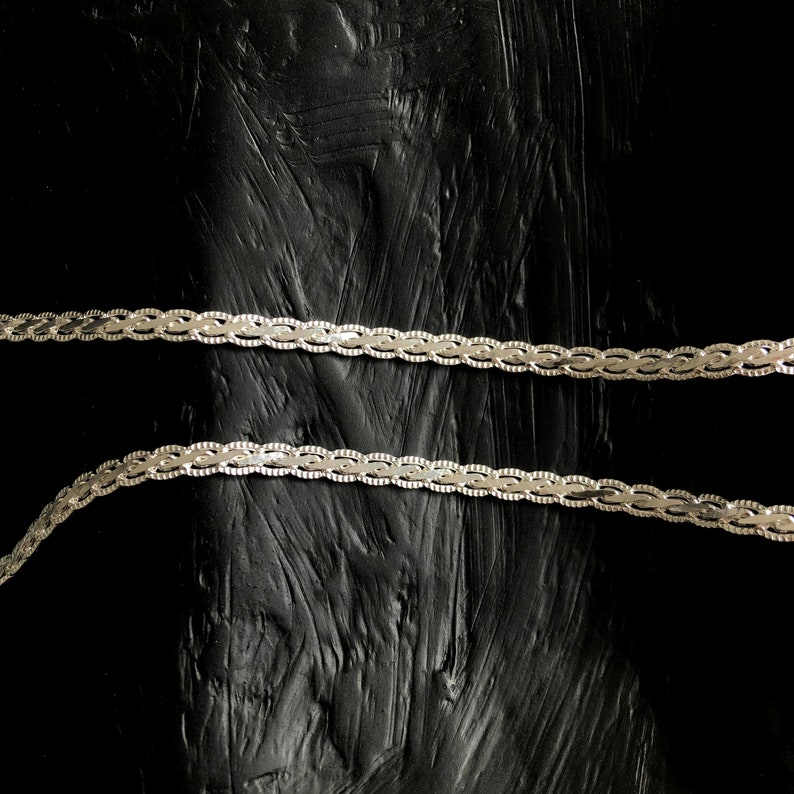 May include: Two silver decorative chains with a repeating oval and swirl pattern. The chains are positioned horizontally against a textured black background. The chains appear to be made of metal. The background has a rough, uneven texture.