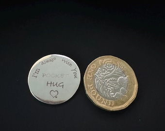 Op de afbeelding: Een zilveren pocket hug munt met de woorden "I'm Always With You POCKET HUG" en een hartsymbool. Daarnaast een goud-zilveren pondmunt.