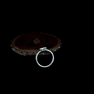 K&ouml;nnte beinhalten: Ein silberner Ring mit einem kleinen, hellfarbigen Edelstein. Der Ring ist vor einem dunklen Hintergrund platziert, mit einem Blick auf eine braune, verzierte Oberfl&auml;che. Der Edelstein scheint herzf&ouml;rmig zu sein.