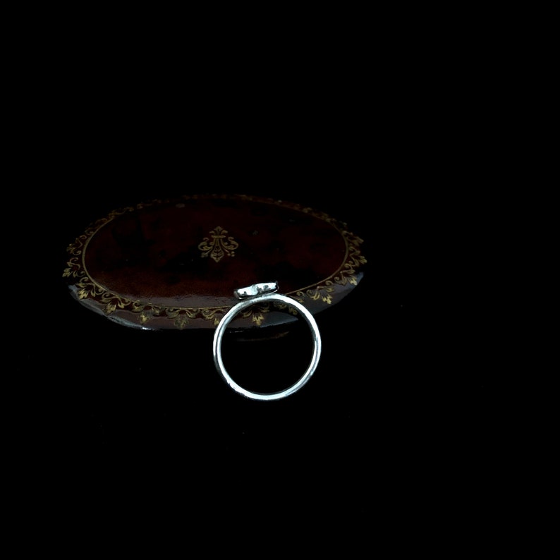 K&ouml;nnte beinhalten: Ein silberner Ring mit einem kleinen, hellfarbigen Edelstein. Der Ring ist vor einem dunklen Hintergrund platziert, mit einem Blick auf eine braune, verzierte Oberfl&auml;che. Der Edelstein scheint herzf&ouml;rmig zu sein.