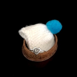 K&ouml;nnte beinhalten: Ein silberner Ring mit herzf&ouml;rmiger Oberfl&auml;che, graviert mit den Buchstaben "LC". Der Ring wird in einem kleinen, braunen, gebogenen Beh&auml;lter pr&auml;sentiert, mit einem wei&szlig;en Strickartikel und einem blauen Pompon im Hintergrund. Der Hintergrund ist schwarz.