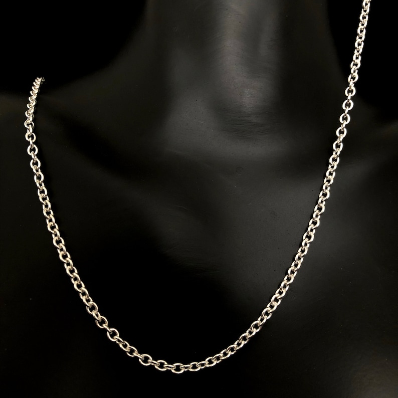 May include: A silver-colored chain necklace with a classic cable link design. The necklace is displayed on a black mannequin, showcasing its simple elegance. The chain is made of a shiny metal.