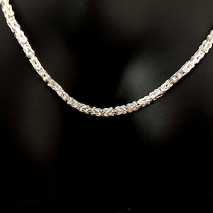 May include: A silver Byzantine chain necklace against a black backdrop. The chain, made of interlocking links, has a textured appearance. The necklace is displayed in a curved shape, showcasing its design.