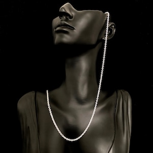 May include: A silver-colored chain necklace displayed on a black mannequin. The chain is a delicate, linked design, hanging from the mannequin's ear and resting on its chest. The background is a solid black.