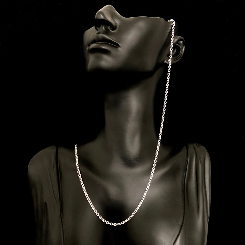 May include: A silver-colored chain necklace displayed on a black mannequin. The chain is a delicate, linked design, hanging from the mannequin's ear and resting on its chest. The background is a solid black.