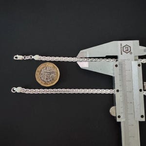 May include: Two silver chain bracelets with a textured design are shown on a black surface. A coin and a measuring caliper are also present. The bracelets have a delicate, woven appearance, and the clasps are visible.