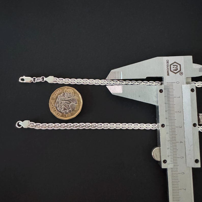 May include: Two silver chain bracelets with a textured design are shown on a black surface. A coin and a measuring caliper are also present. The bracelets have a delicate, woven appearance, and the clasps are visible.