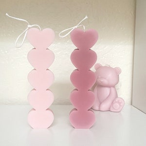 Puede incluir: Dos velas de color rosa en forma de corazón, una rosa claro y otra rosa más oscuro, cada una con cinco corazones apilados uno encima del otro. Las velas están sobre una superficie blanca.
