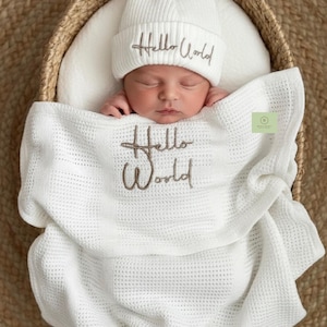 May include: A newborn baby wrapped in a white waffle-knit blanket and wearing a matching beanie. The words "Hello World" are embroidered on both the hat and blanket in a brown thread. The baby is nestled in a woven basket.